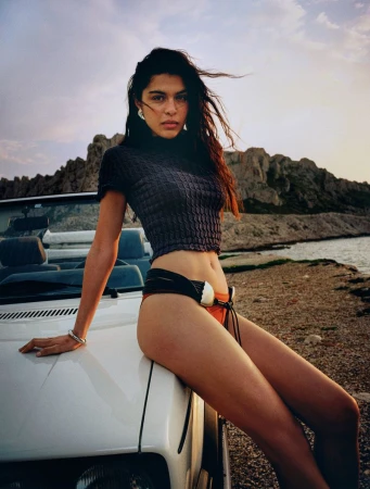 Woman sits on car hood near ocean at dusk, wearing a crop top and bikini bottoms, with rocks and sky in the background.
