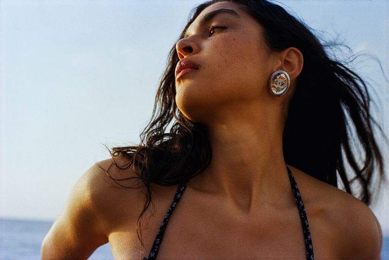 Adult female poses in outdoor setting with view of water and clear blue sky. Woman wears earrings and bikini top, bathed in sunlight.