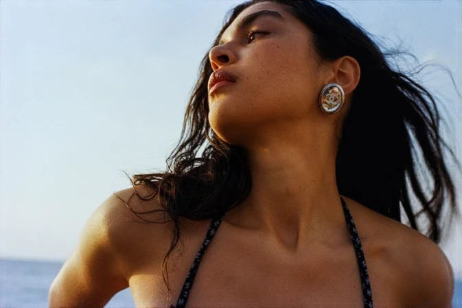 Adult female poses in outdoor setting with view of water and clear blue sky. Woman wears earrings and bikini top, bathed in sunlight.