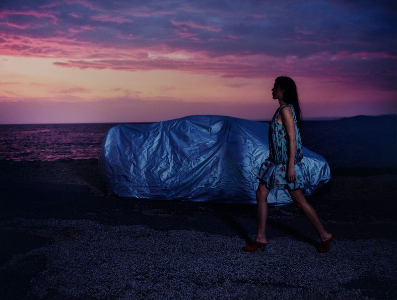 A woman in a floral dress walks along a rocky beach past a covered car at dusk, with a soft sunset in the background.