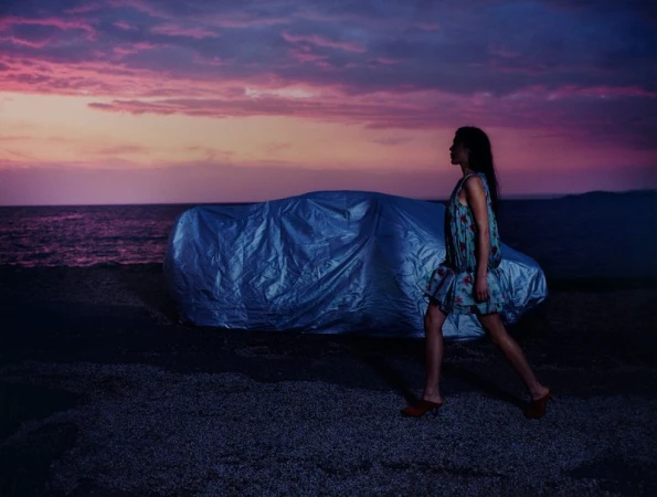 A woman in a floral dress walks along a rocky beach past a covered car at dusk, with a soft sunset in the background.