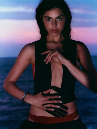 Woman with hands posed on waist in an unbuttoned black vest against twilight seascape backdrop. Jewelry, red accents in natural light.