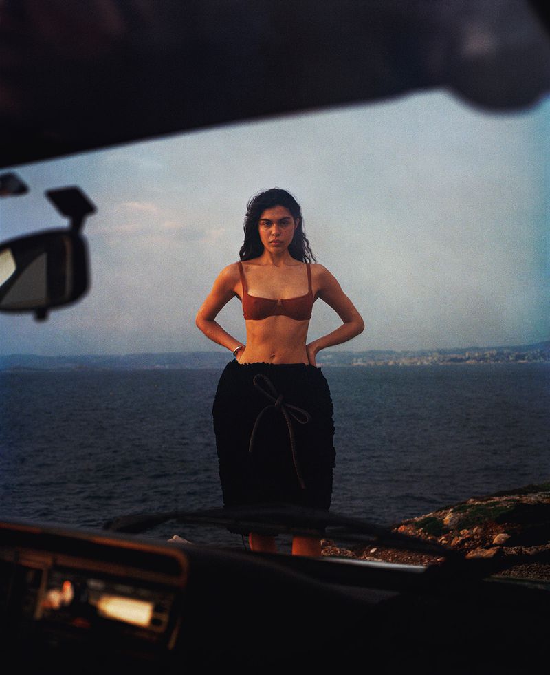 Woman in a brown bikini top and black skirt standing near the ocean, with a car interior visible in the foreground.