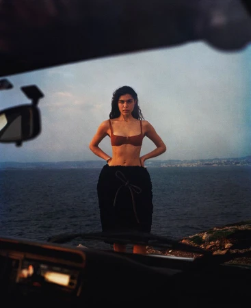 Woman in a brown bikini top and black skirt standing near the ocean, with a car interior visible in the foreground.