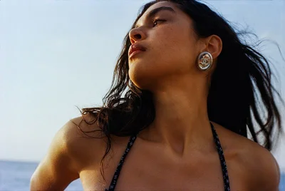 Close-up portrait of a woman with dark hair blowing in the wind wearing earrings and a patterned strap top against an outdoor sky background.