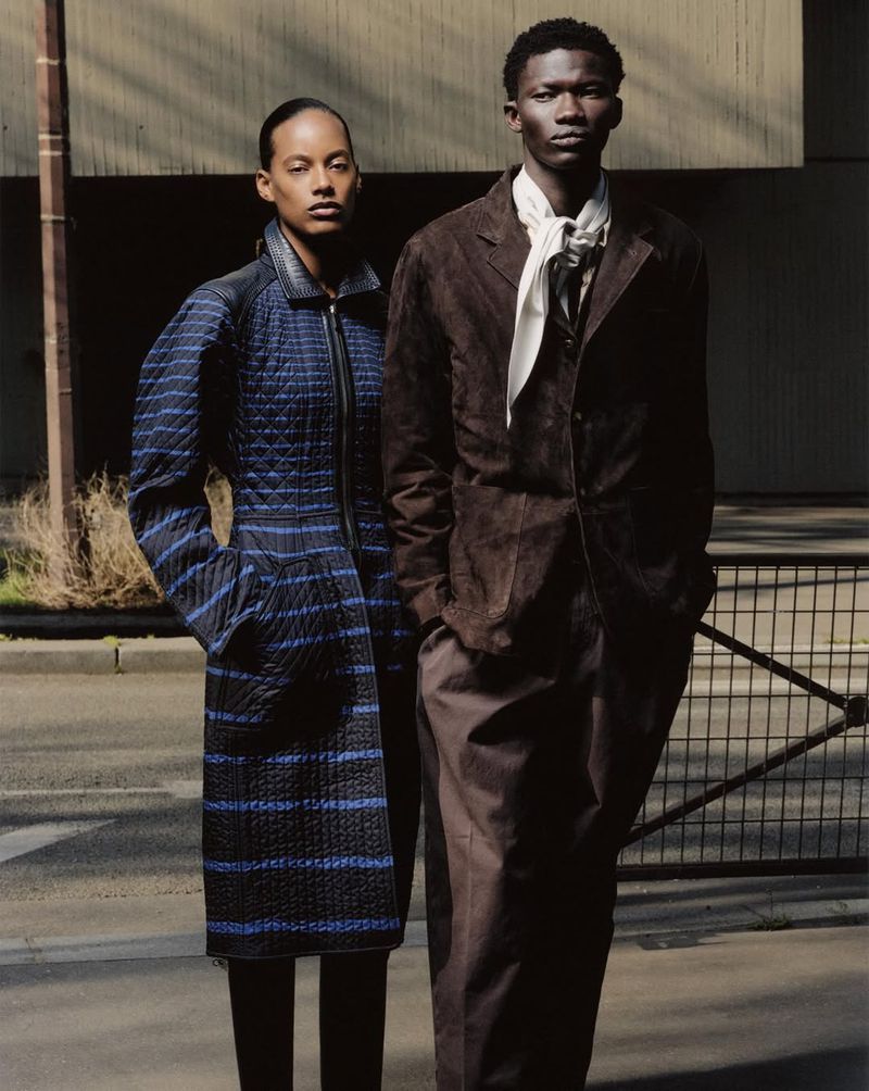 Two fashion models posing outdoors, one in a quilted striped blue coat and the other in a brown suede jacket with a tied white scarf.