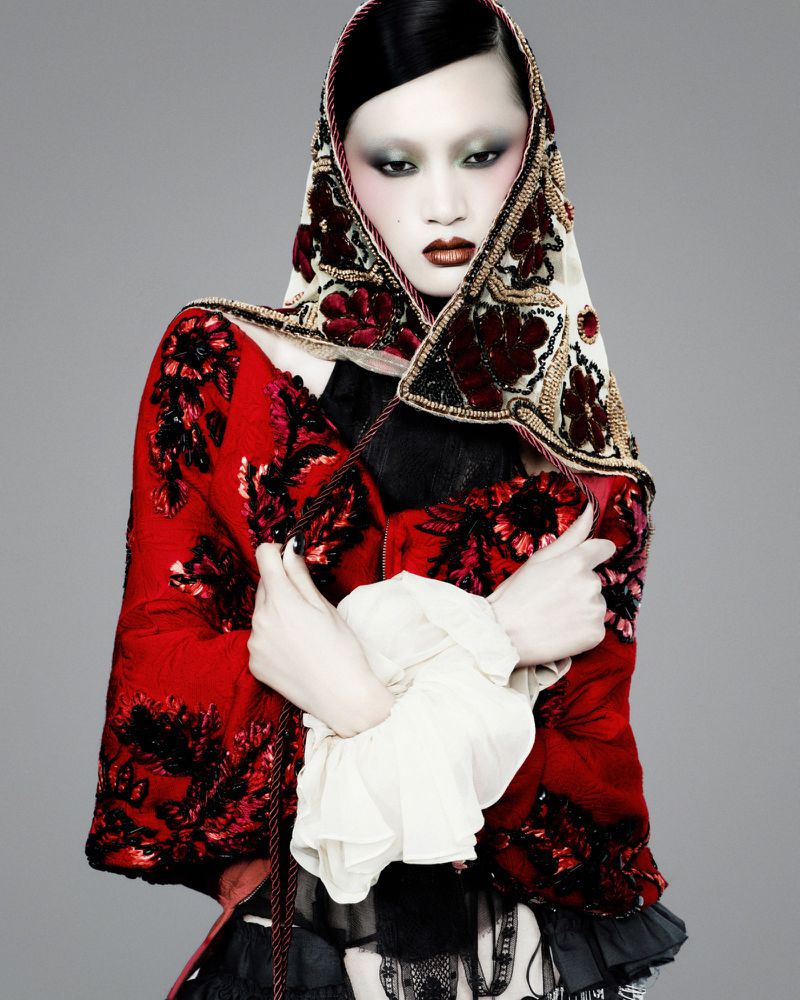 Model Yilan Hua wearing an embroidered red cape and floral headscarf against a grey studio backdrop.