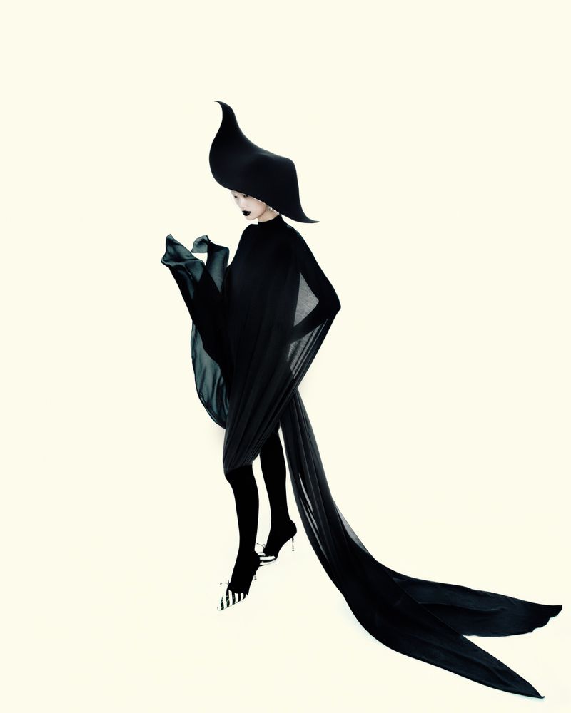 Model Yilan Hua wearing an avant-garde black gown, sculptural hat, and striped heels against a pale seamless studio background.