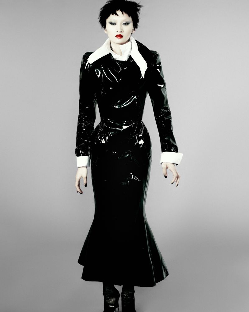 Model Yilan Hua wearing a glossy black vinyl dress with a white collar against a grey studio backdrop.
