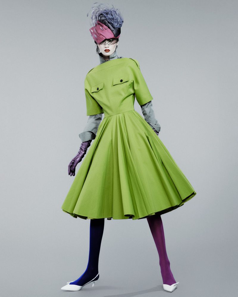 Model Yilan Hua posing in a lime green pleated dress with an avant-garde fascinator hat against a grey seamless background.