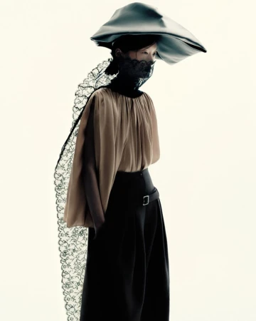 Model Yilan Hua wearing a sculptural hat, black lace veil, pleated beige blouse, and high-waisted black trousers on a white backdrop.