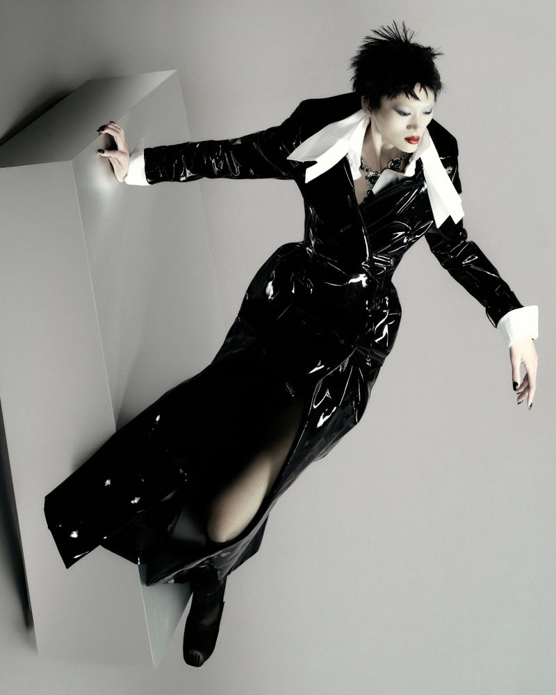 Model Yilan Hua in a glossy black vinyl coat with an oversized white collar leaning over a grey geometric plinth.