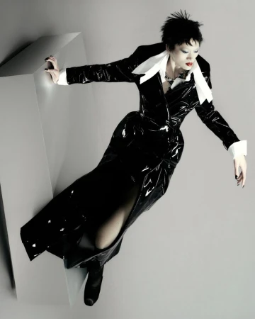 Model Yilan Hua in a glossy black vinyl coat with an oversized white collar leaning over a grey geometric plinth.