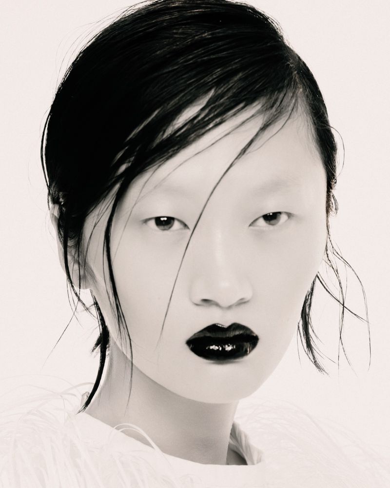 Close-up beauty portrait of model Yilan Hua with slicked wet hair and glossy black lipstick against a white seamless background.
