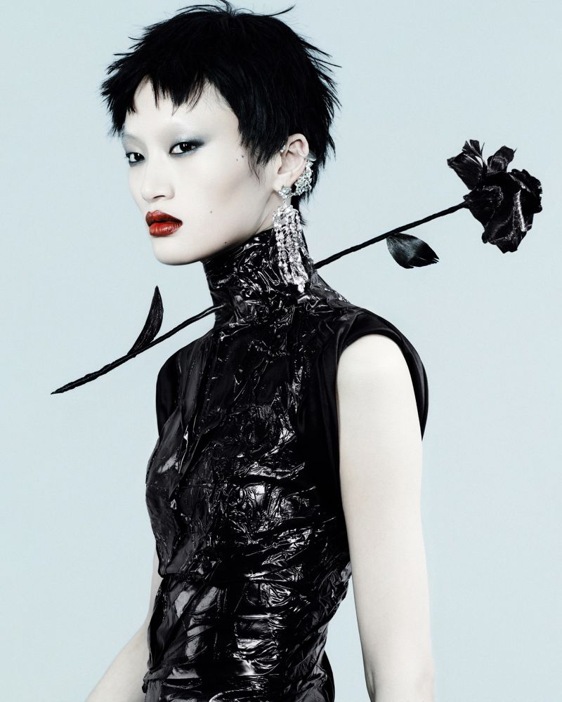 Model Yilan Hua with short black hair in a glossy high-neck black top with a black rose through the collar on a light blue background.