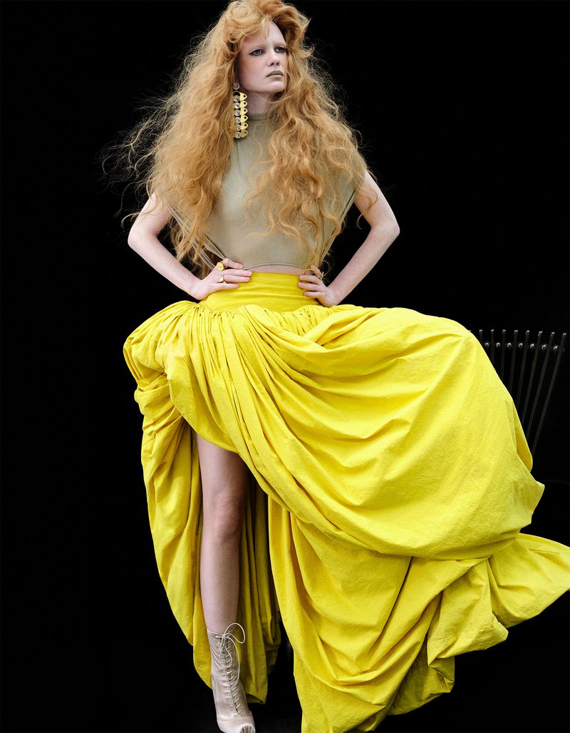 Fashion model with voluminous curly red hair posing in a dramatic draped yellow maxi skirt and beige top against a black backdrop.