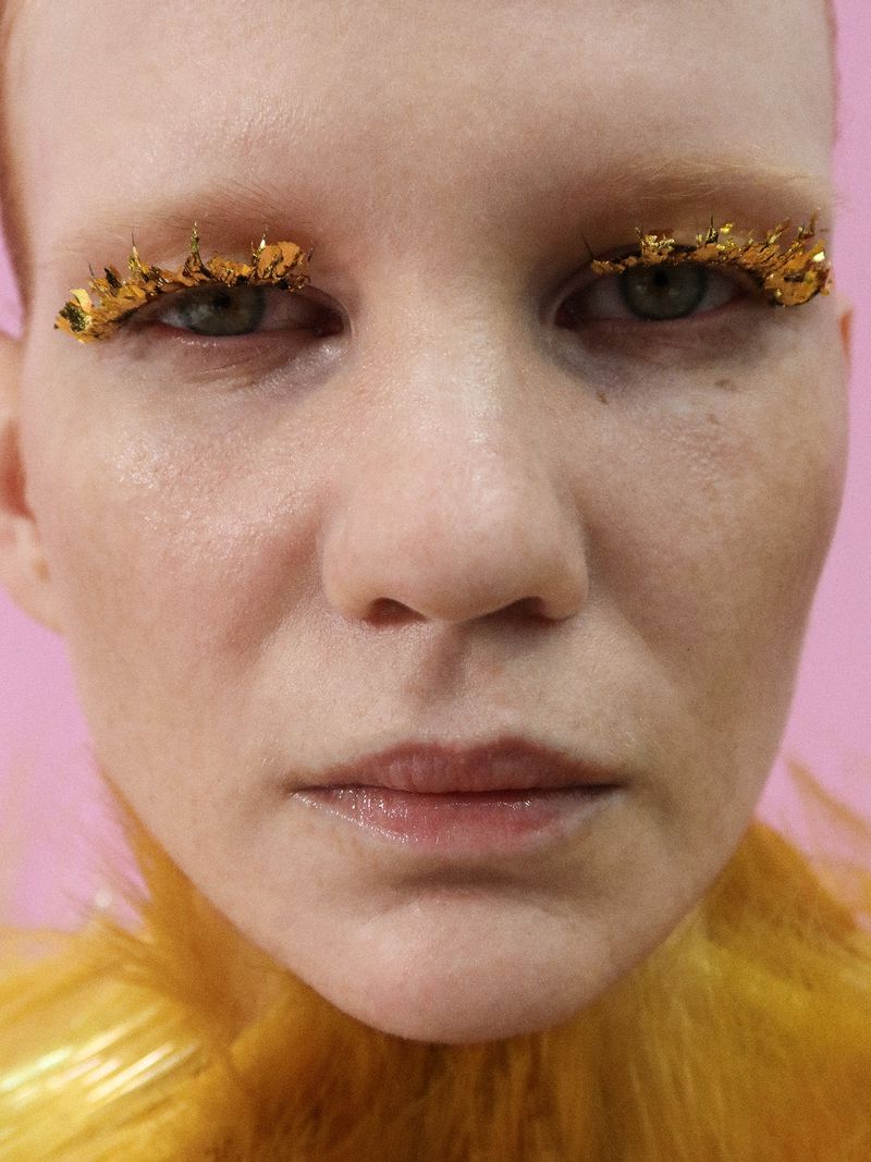 Extreme close-up beauty portrait of a model with gold leaf eyelashes wearing a yellow feather collar against a pink backdrop.