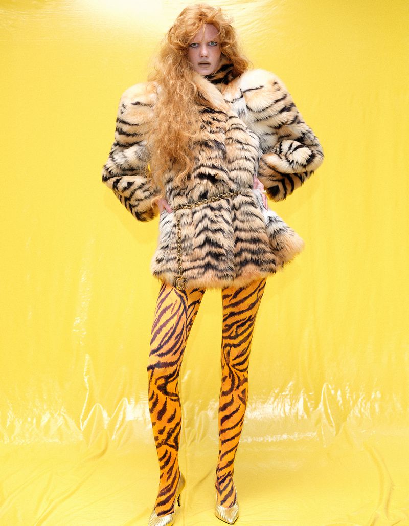 Fashion model with voluminous crimped hair wearing a tiger-print fur coat and tiger tights against a vibrant yellow backdrop.