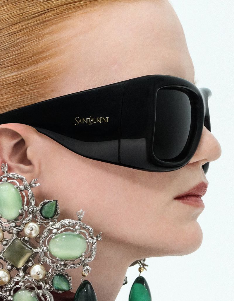 Close-up profile of a woman with red hair wearing oversized black Saint Laurent sunglasses and ornate green gemstone drop earrings.