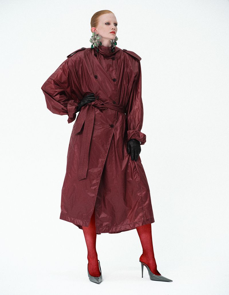 Model posing in an oversized burgundy belted trench coat, red stockings, grey pointed heels, and statement earrings on a white seamless backdrop.