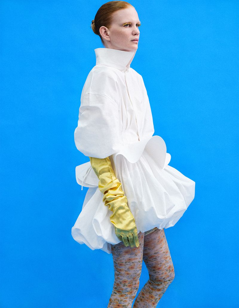 Model in a voluminous white dress and yellow satin gloves standing against a bright blue studio backdrop.