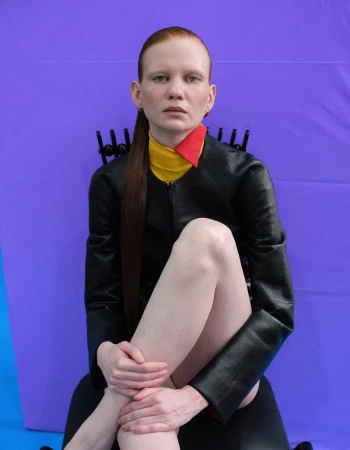 Model with slicked red hair seated on a black chair wearing a black leather jacket against a vivid purple backdrop.