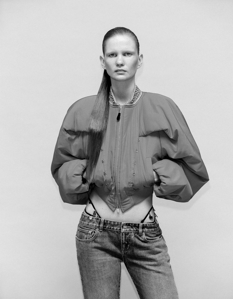 Model wearing a structured cropped bomber jacket and low-rise denim jeans against a plain studio background in black and white.