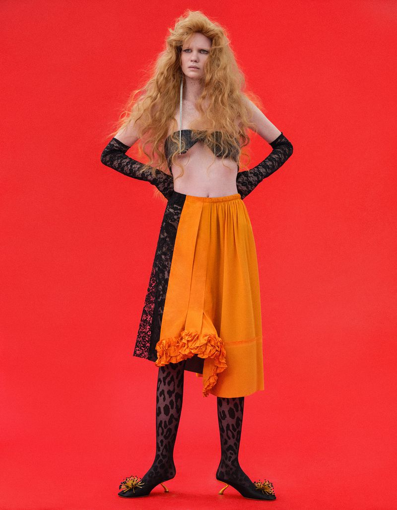 Fashion model in an orange ruffled skirt, black lace gloves, and leopard print tights against a vibrant red backdrop.