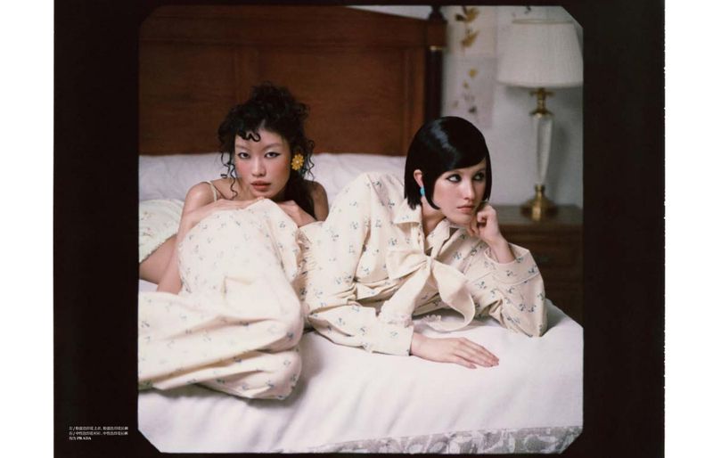 Two women in floral printed outfits rest across a white-sheeted bed in front of a wooden headboard and classic bedside lamp.
