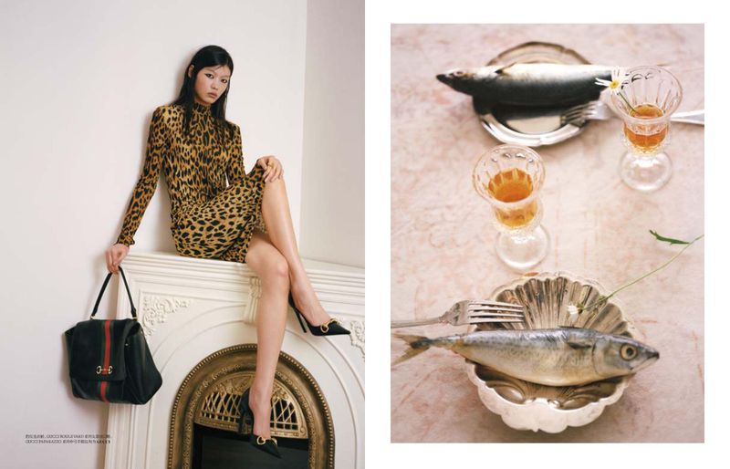 Model Tang He in a leopard print dress seated on a white fireplace mantel alongside a still life of fish and glassware on marble.