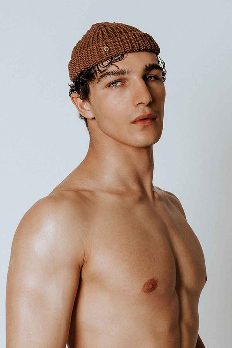 Close-up studio portrait of a shirtless adult male wearing a brown beanie hat and looking at the camera with a neutral expression.
