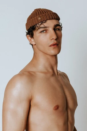Close-up studio portrait of a shirtless adult male wearing a brown beanie hat and looking at the camera with a neutral expression.