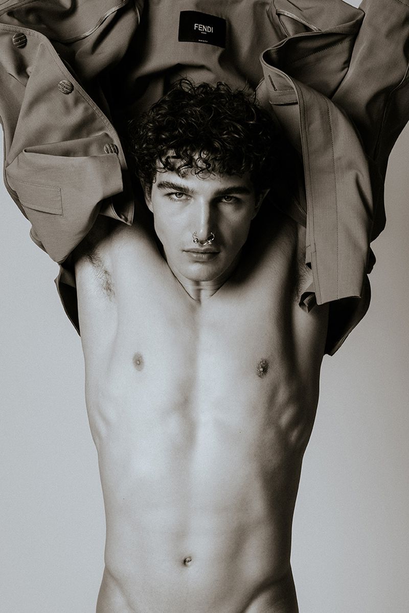 Portrait of a shirtless adult man with curly hair, a nose ring, and a partially removed coat, against a neutral backdrop in a studio.