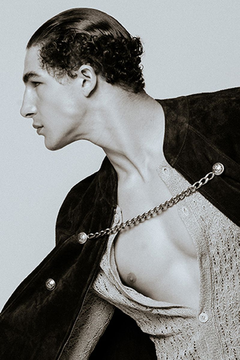 Monochrome portrait of a male model with slicked-back hair wearing a light-colored knitted shirt and a dark jacket with a silver chain.