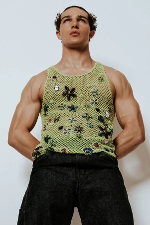 Model Sacha Bilal wearing an embellished lime green crochet tank top and black jeans against a white seamless background.