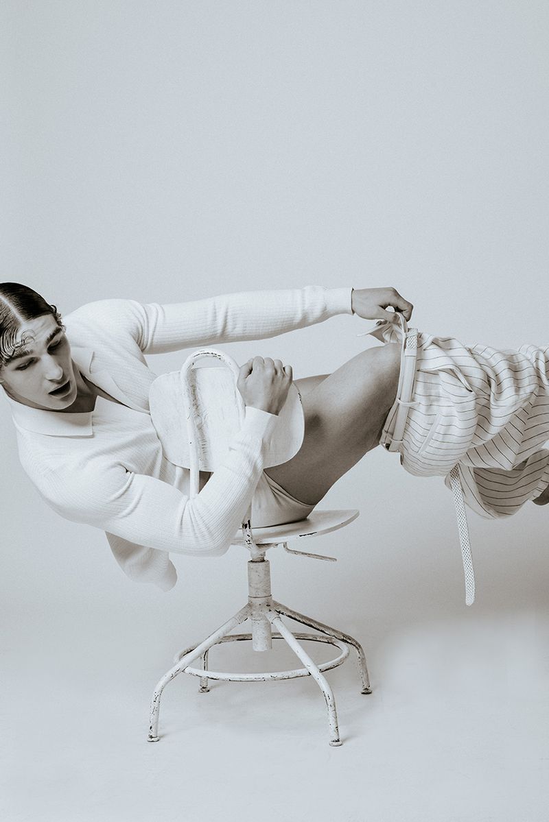 Black-and-white portrait of a slender adult male lounging on a white chair, wearing a long-sleeve shirt and striped pajama bottoms.
