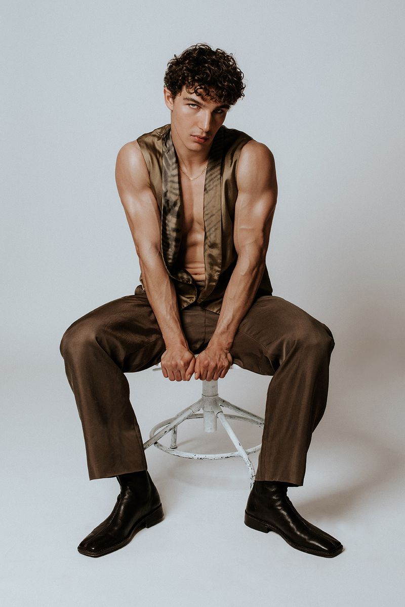 Attractive male fashion model sits on a stool wearing brown silk top and trousers with black leather boots in a neutral studio setting.