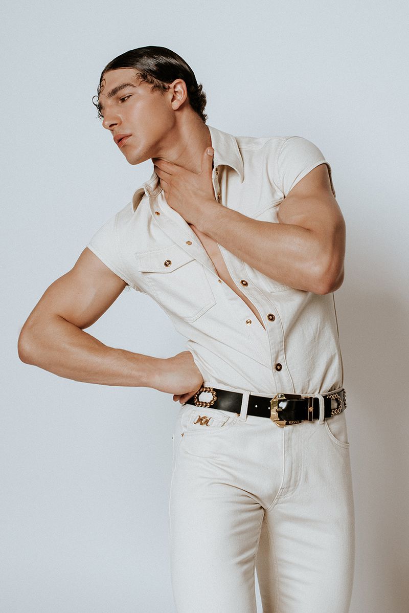 Portrait of a slender adult man in a sleeveless cream button-down shirt and cream pants, posing in front of a pale background.