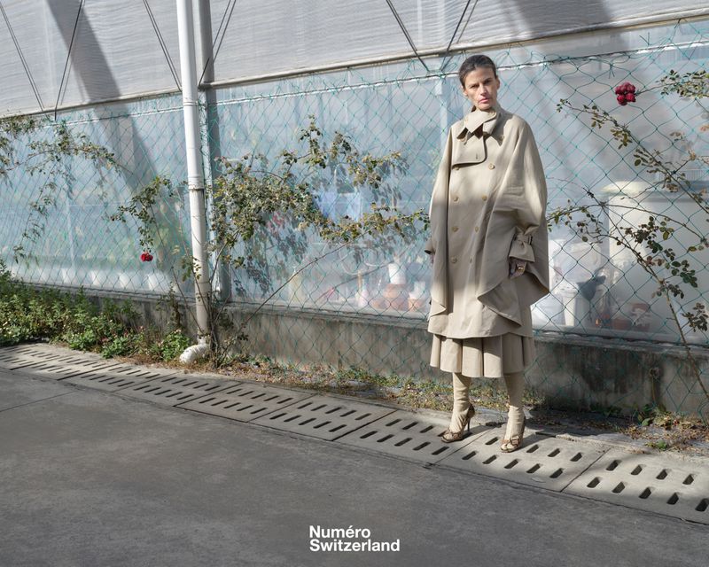 Model wearing an oversized beige cape trench coat standing by a greenhouse wall with rose bushes.