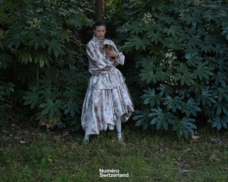 Woman in a voluminous floral brocade gown holding a cat while standing surrounded by lush green foliage.