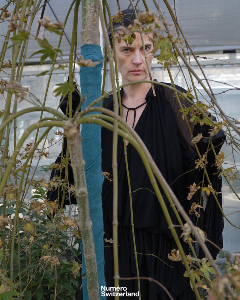 Model in black draped clothing gazing forward through thin tree branches inside a greenhouse for Numéro Switzerland.