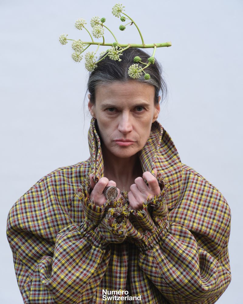 Model in a high-collared plaid coat with a botanical branch balanced on her head against a plain pale grey background.