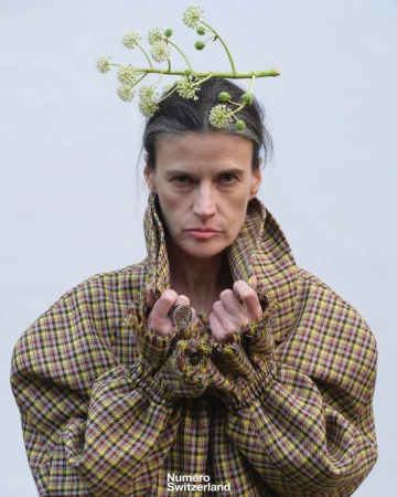 Model in a high-collared plaid coat with a botanical branch balanced on her head against a plain pale grey background.