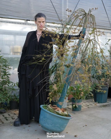 Model wearing a draped black gown posing next to a tall potted weeping tree inside a plant nursery greenhouse.