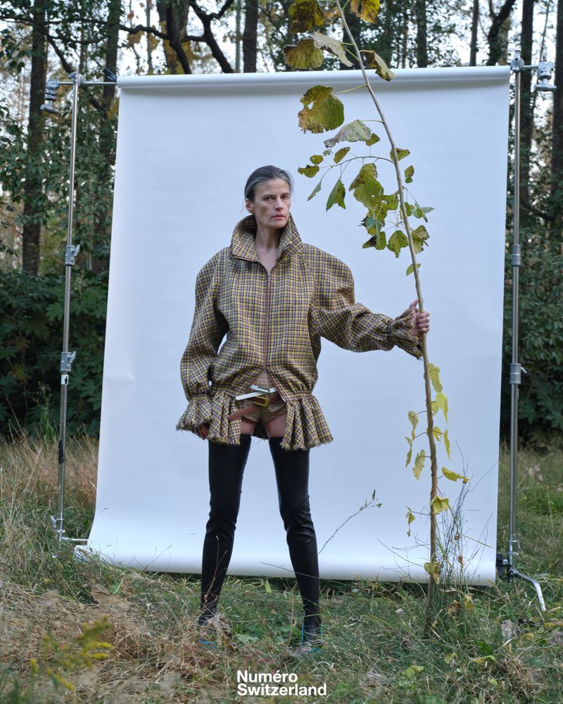 Model wearing a plaid ruffled jacket and black thigh-high boots holding a tall leafy branch before an outdoor white backdrop.