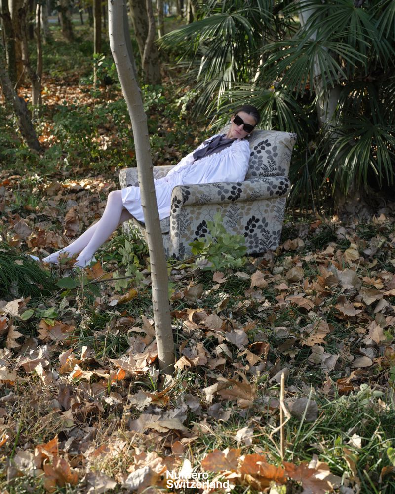 A woman wearing a white dress, white tights, and dark sunglasses reclines in a patterned armchair outdoors among fallen autumn leaves.