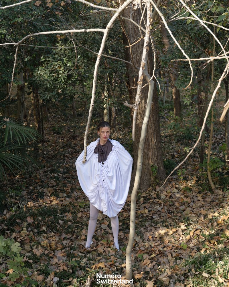 Model in a voluminous white draped garment standing among fallen leaves in a forest under natural light