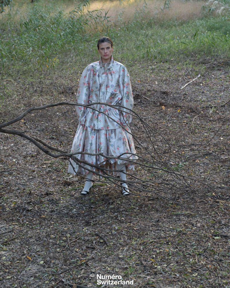 Model standing outdoors behind bare tree branches in a voluminous silver floral tiered coat dress for Numéro Switzerland.