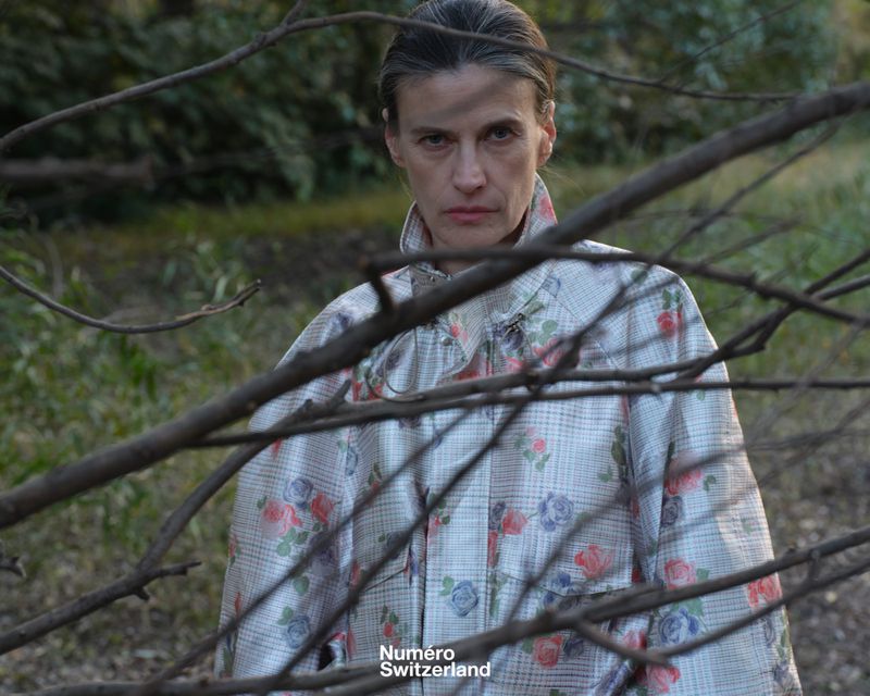 Woman in a floral patterned jacket looking at the camera through bare tree branches in an outdoor garden setting.