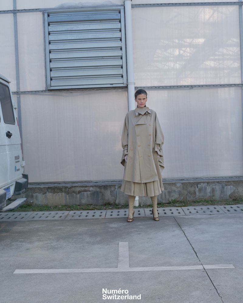 Model posing in an oversized beige layered trench coat and pleated skirt in front of a pale industrial wall.